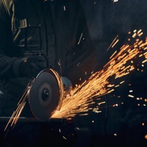 Close up of man in special dark suit and black protective gloves working angle grinder for metal with large flash sparks. Concept of process working electric steel cutter machine.